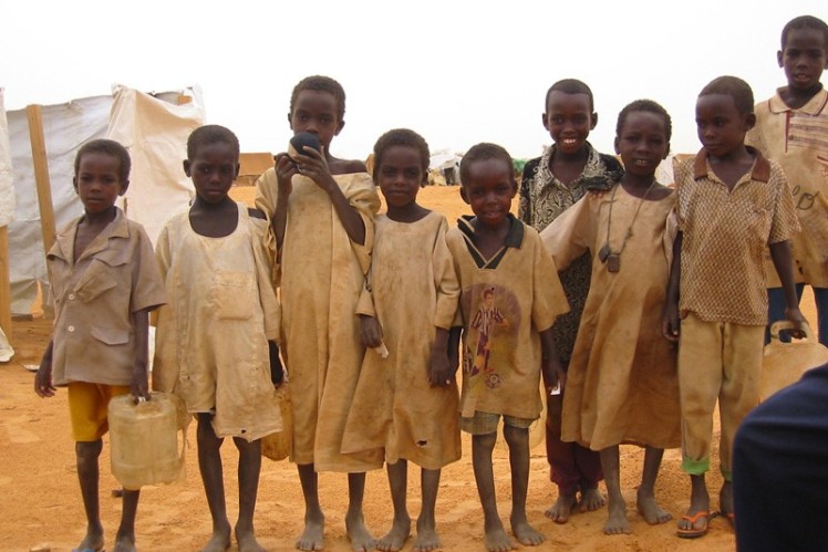 Refugee children stand in a row. A few children are holding a water canister in their hands.