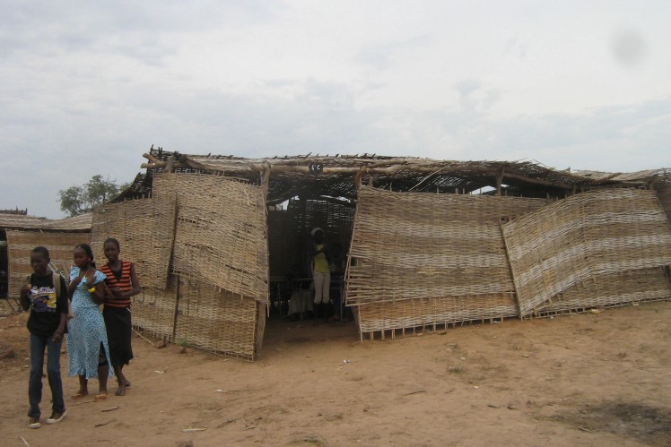 Three girls stand at the entrance to their classroom, which is made of straw.