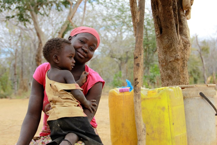 Woman sits next to a yellow water canister and has her child on her lap.