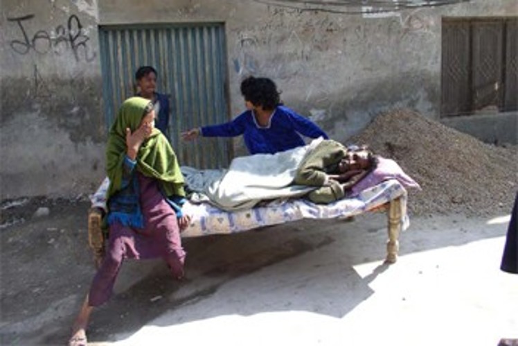 A sick man lies in bed in the middle of the road, while his wife sits next to him, crying.
