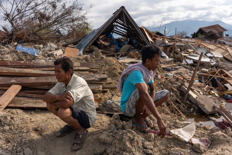 Two men sit in front of a destroyed settlement.