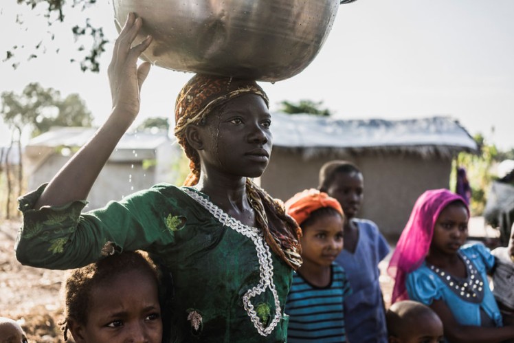 Young girl carries a metal bowl of water on her head.